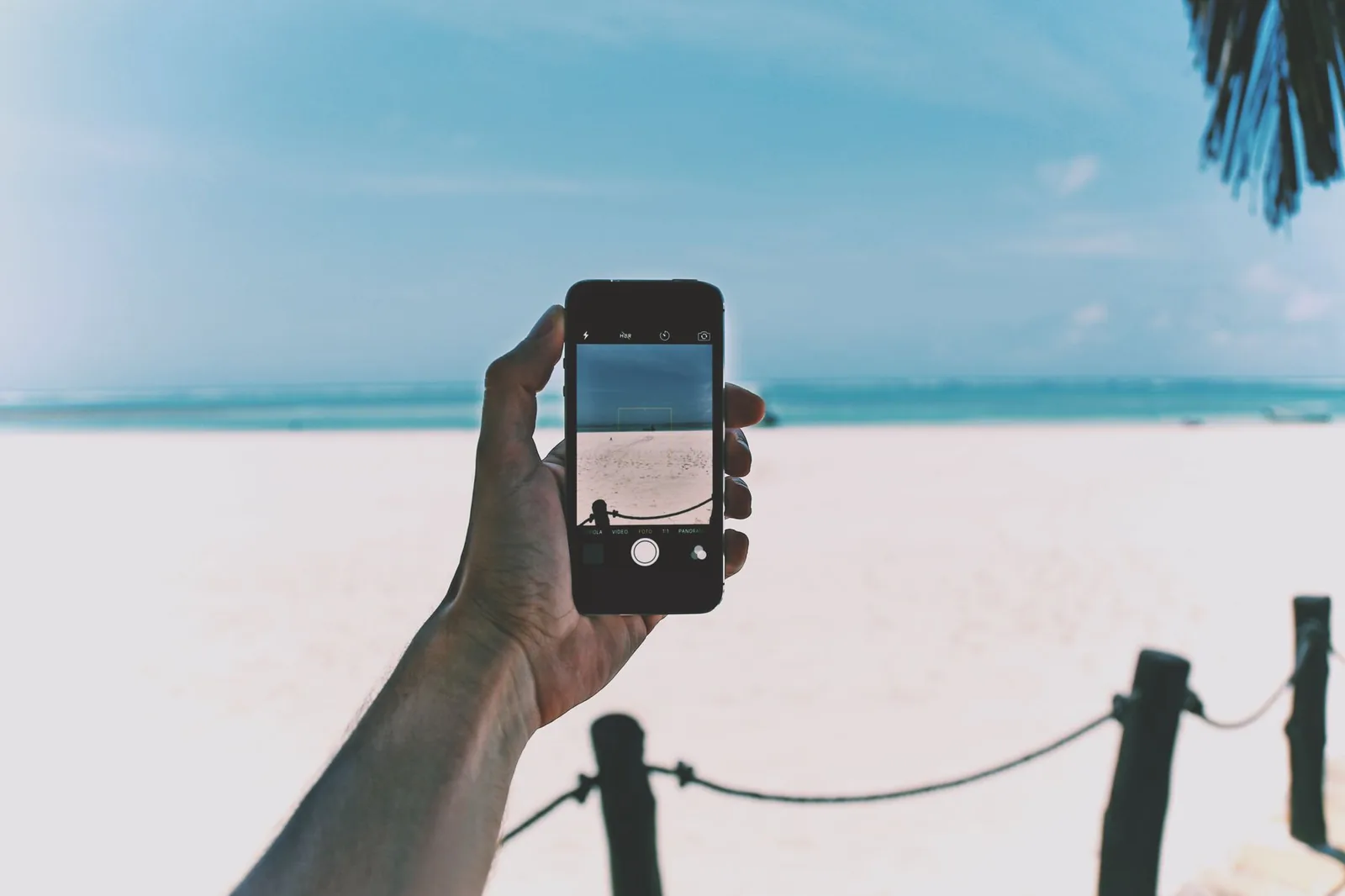 A hand holding a phone up against the white sand and turquoise water of a Zanzibar beach
