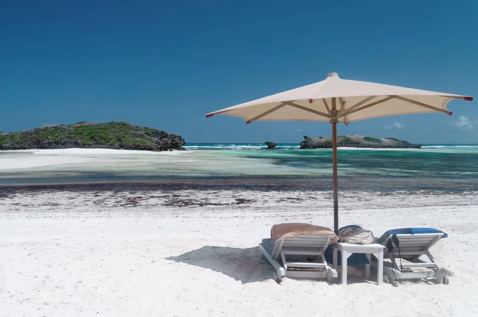 Sun loungers and a parasol on white sand facing a turquoise lagoon and rocky islets on the Indian Ocean