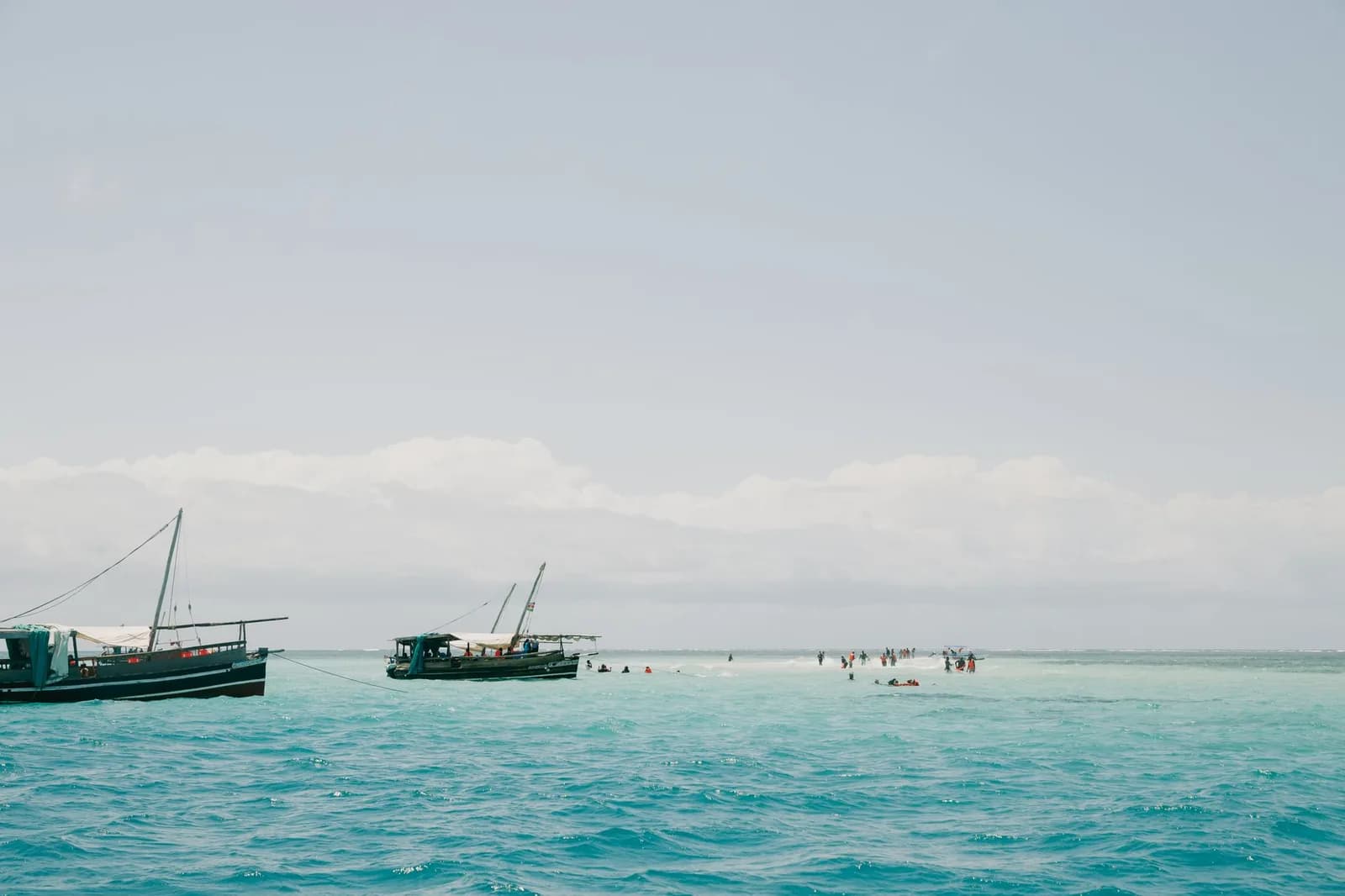 Two wooden dhows moored in turquoise water beside a sandbar where a group of tourists stands and swims
