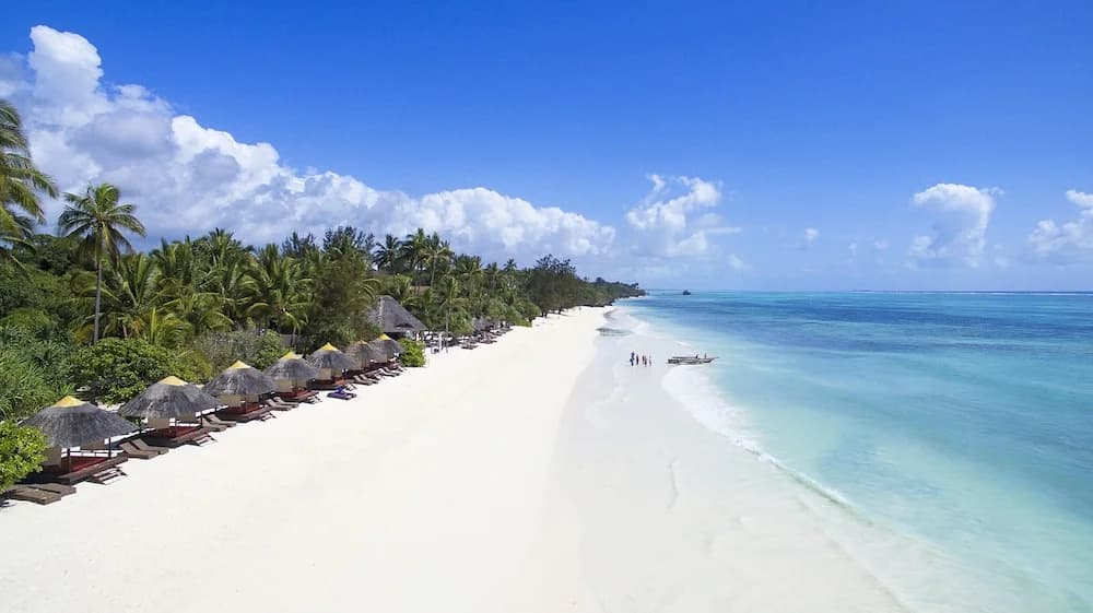 Aerial view of the white sand beach at Meliá Zanzibar in Kiwengwa, with thatched parasols and loungers along the palm-lined shore and a turquoise lagoon