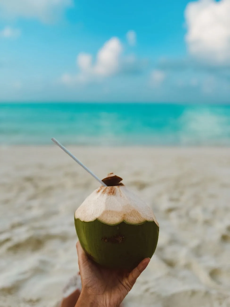 A green drinking coconut with a straw held up on a white sand beach in Zanzibar