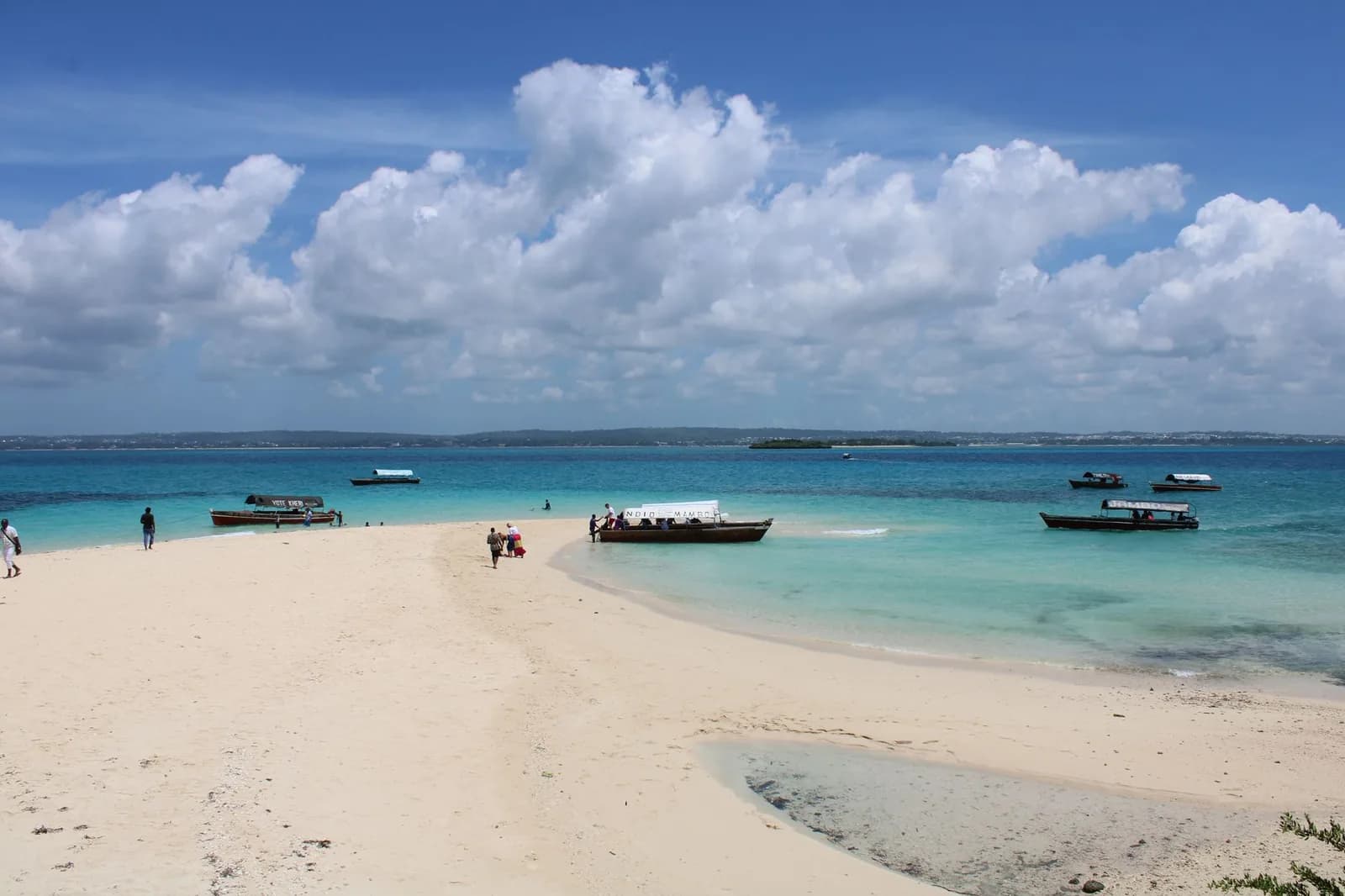 White sandbar emerging from shallow turquoise water with wooden boats anchored around it and a few visitors on the sand, Zanzibar