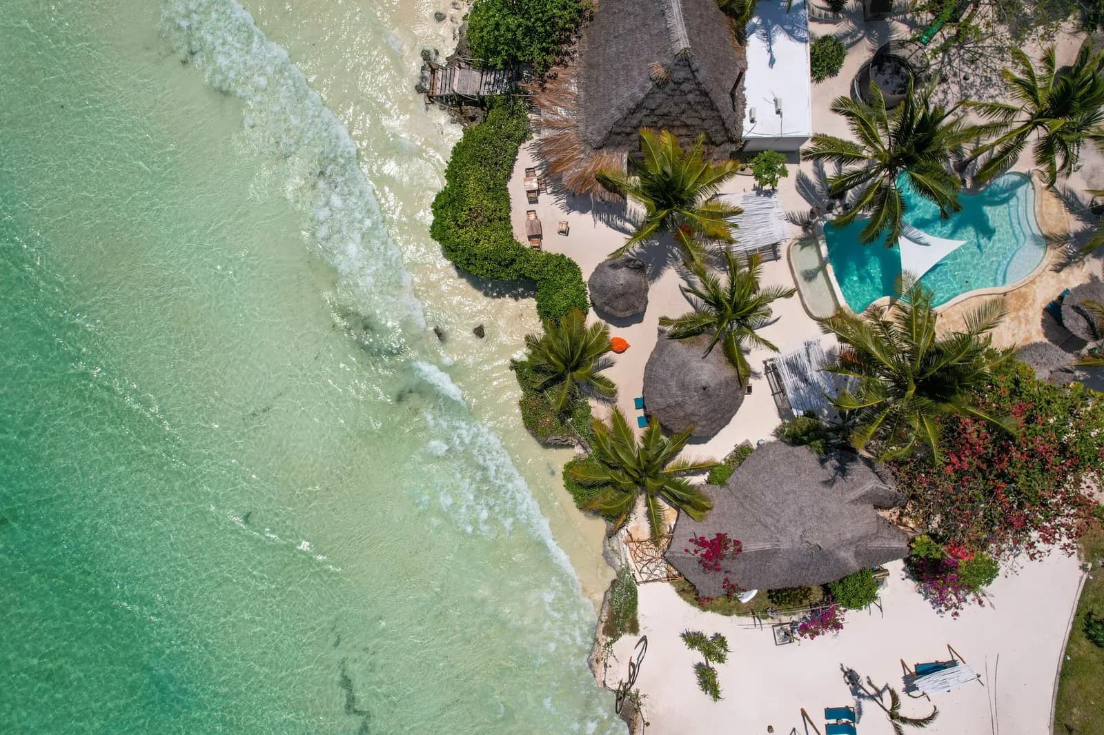 Thatched roofs, palms and a resort pool seen from above, next to the turquoise water of a Zanzibar lagoon