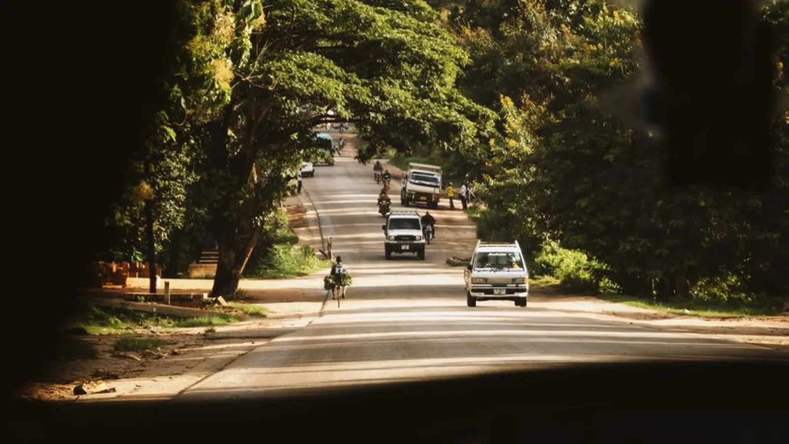 A Zanzibar road seen from inside a taxi, with a dala dala and cyclists under the trees