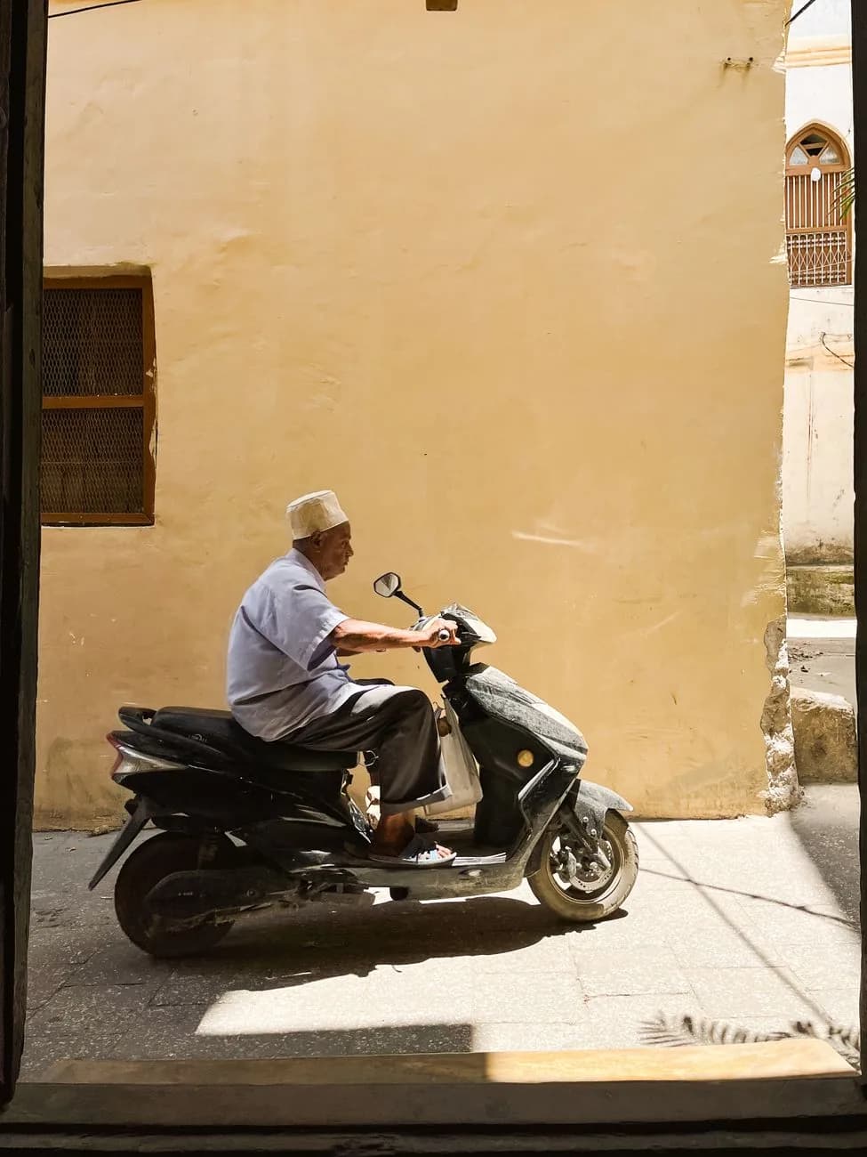 A man in a white kofia riding a scooter through a sunlit lane in Stone Town, Zanzibar